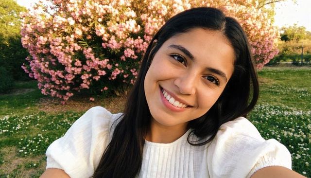 Smiling woman taking candid selfie in sunlit garden with pink blossoms at golden hour