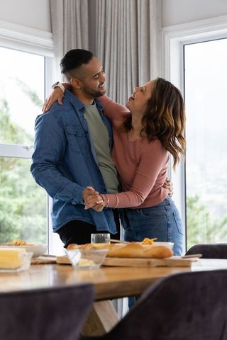 Mid-adult couple dancing and embracing by sunlit kitchen table with fresh bread