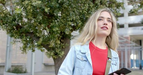 Young woman holding smartphone in urban tree-lined walkway wearing denim jacket, red top