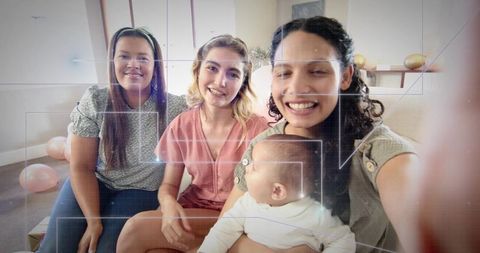 Smiling friends taking selfie with infant in cozy pastel living room at small celebration