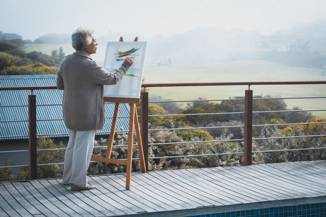 Senior African American woman painting on a canvas while standing on a balcony at home. Ideal for use in articles or advertisements related to senior lifestyle, retirement activities, creative hobbies, and the benefits of art for mental health. Can also be used to depict peaceful and relaxing home environments.