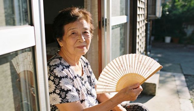 Senior woman sitting on doorstep holding decorative folding fan in sunlit doorway