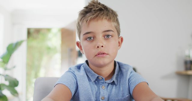 Young boy sitting at indoor table during daytime with a neutral expression, conveying a thoughtful mood. Suitable for educational content, child-centric advertising, psychological well-being topics, lifestyle blogs, or modern family settings.
