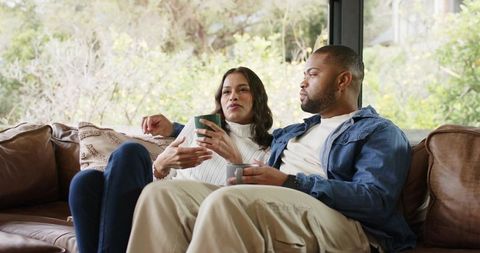 Diverse couple relaxing on leather sofa holding mugs and talking in bright living room