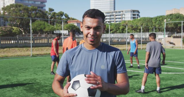 Young male soccer player holding a ball, smiling on outdoor field with teammates, wearing athletic uniform. Ideal for content related to sports, team training sessions, youth athletic activities, and outdoor exercises.