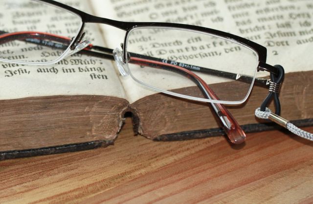 Reading glasses resting on open vintage book with aged pages on a wooden surface. This image can be used to represent studying, literature, acquiring knowledge, or a love for reading. Suitable for educational or literary themes, and ideal for use in articles, blogs, or websites focused on education, books, or vintage items.