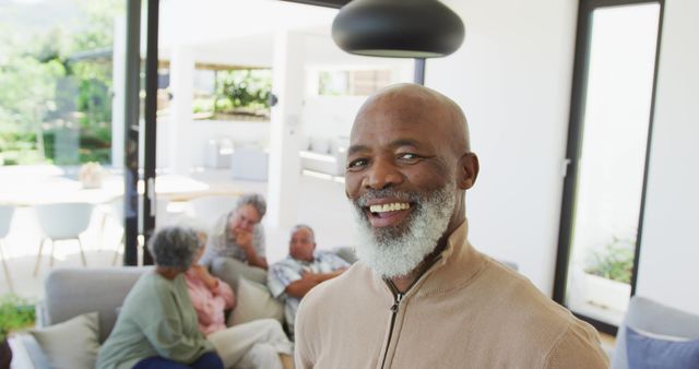 Senior man is smiling warmly while standing in a modern well-lit living room. In the background, family members of different generations are engaged in conversations, illustrating togetherness and familial bond. This image is ideal for advertisements for retirement communities, family-centric services, or happiness in later stages of life.