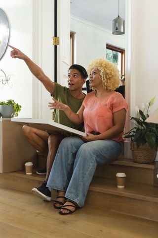 Multiracial couple sitting on steps holding artwork and pointing while moving into home