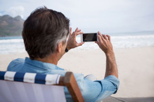 Man sitting on beach chair, capturing scenic view of ocean with mobile phone. Ideal for travel blogs, vacation advertisements, technology use in nature, and lifestyle articles.