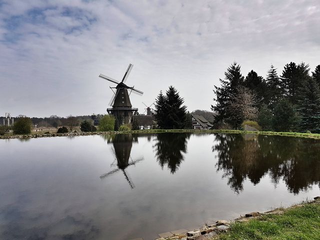Traditional windmill reflecting on calm pond on a partially cloudy day with blue sky and surrounding trees. Ideal for use in travel brochures, environmental websites, and digital wallpapers showcasing serene rural landscapes.