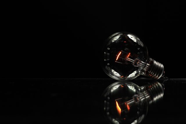 This stock photo features a close-up of an illuminated lightbulb lying on a reflective surface in a dark room. The glowing filament and dark background create a dramatic contrast, making it ideal for technology, innovation, or energy-related projects. It can be used in presentations, advertisements, or educational materials to symbolize enlightenment, bright ideas, or modern technology.