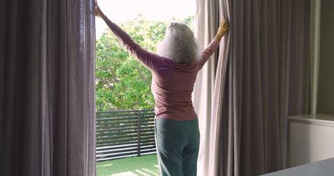 Senior Woman Opening Curtains for Morning Sunlight on Balcony