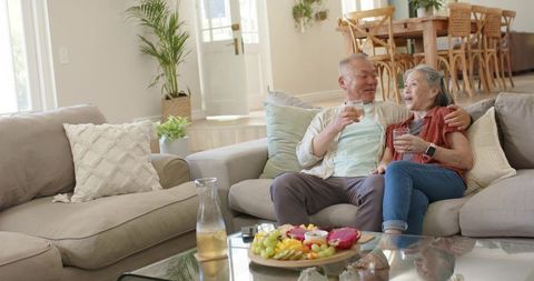 Senior asian couple relaxing on gray sofa sharing fresh fruit and drinks in bright living room