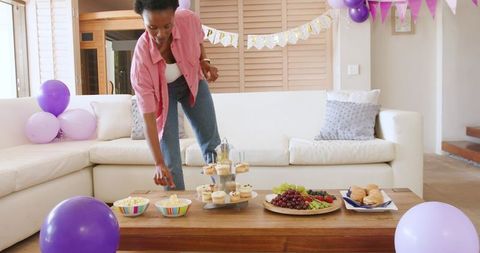 African american woman hosting pastel living room birthday, arranging cupcakes and snacks