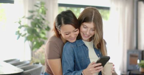 Diverse female couple hugging and looking at smartphone while holding mug in bright home