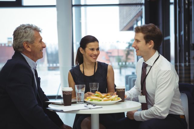 Business colleagues are sitting around a table, engaging in a friendly discussion while having breakfast. They are enjoying coffee and sandwiches, indicating a casual yet professional setting. This image is ideal for illustrating concepts related to teamwork, corporate culture, business meetings, and professional interactions.