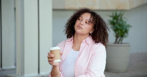 Pensive african american woman holding takeaway coffee in pastel courtyard with greenery