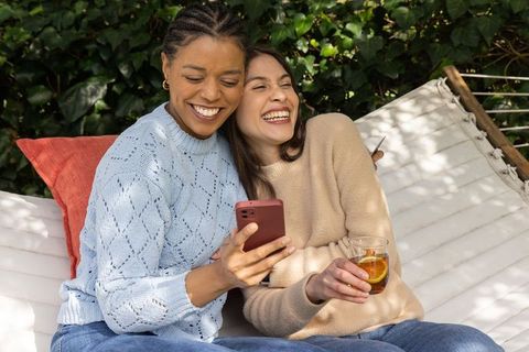 Two women laughing on hammock, sharing smartphone and drink in sunlit cozy backyard lounge