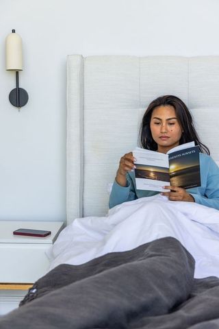 Asian woman reading paperback in bed relaxing in minimalist neutral bedroom calm morning
