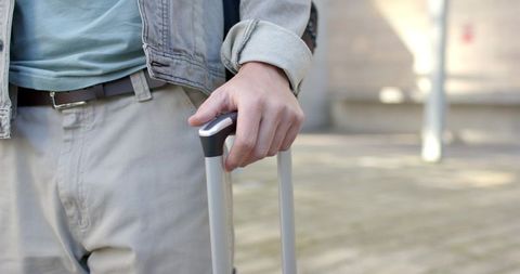 Male traveler gripping suitcase handle walking urban plaza wearing denim jacket