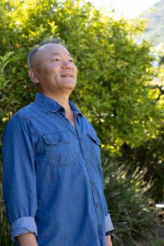 Senior asian man gazing upward in sunlit garden wearing denim shirt and wristwatch