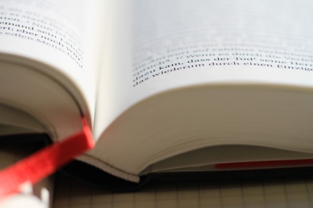 Close-up of an open book with a red ribbon bookmark seen laid across the center. The focus is on the book pages and the text is partially visible, adding an element of mystery about the content. Ideal for use in materials related to studying, reading, literature promotion, educational projects, or library advertisements.