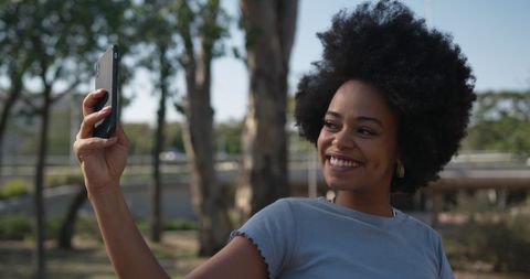 A cheerful biracial woman with curly hair capturing a selfie in a sunlit park. Perfect for content about joyous moments, outdoor lifestyles, or technology in everyday settings. Suitable for ads, web content, or articles celebrating diversity and natural beauty.