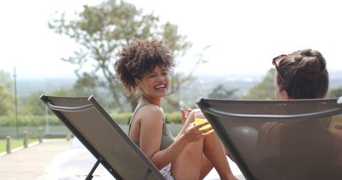 Laughing friends relaxing on pool deck loungers holding refreshing summer drinks