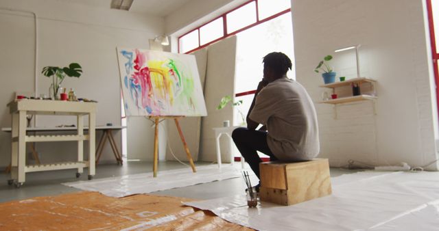 Artist is admiring a colorful abstract painting on an easel in their studio. The workspace is filled with creative tools and supplies, suggesting a dedicated environment for artistic expression. Use for concepts of contemplation, creativity, and the artistic process.