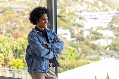 African american man leaning at floor-to-ceiling window in modern home wearing denim jacket