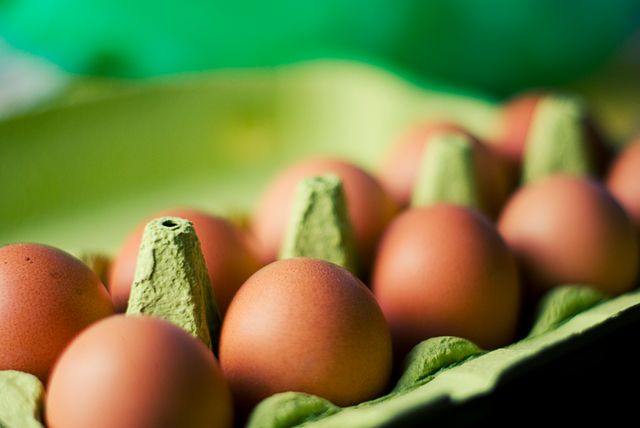 Close-up of brown eggs neatly arranged in a green carton with a blurred green background. This image is ideal for use in content related to grocery shopping, organic food, culinary ingredients, or promoting fresh produce. The vibrant color and focus on eggs can add a warm and natural aesthetic to food blogs, cooking websites, and health-related articles.