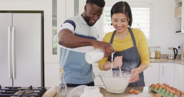 Image depicts a cheerful ethnically diverse couple baking together in a kitchen, both wearing aprons, suggesting collaboration and affection. Ideal for content emphasizing healthy relationships, home cooking blogs, family bonding activities, and lifestyle choices. Useful for articles, ads, and social media posts focusing on spending quality time at home, modern family life, and the joy of cooking together.