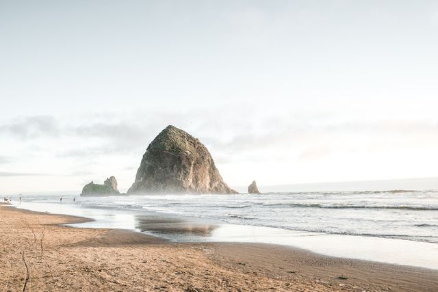 This serene coastal scene captures a towering rock formation rising from the ocean near a peaceful sandy shore. Ideal for travel blogs, beach destination guides, nature websites, and background images in presentations.