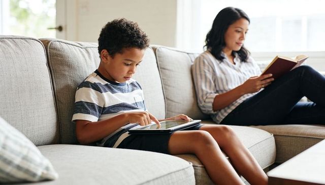 Young boy tapping tablet on couch while parent reading book in bright living room