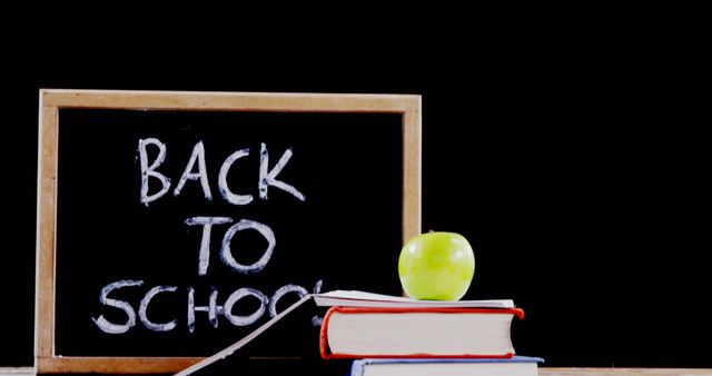 Chalkboard with 'Back to School' message written in white chalk, standing on desk alongside stacked books and green apple, suggesting fresh start of new academic year. Ideal for educational blogs, school promotions, and classroom decorations.