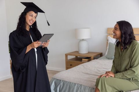 Graduate wearing cap and gown celebrating with friend at home using tablet, virtual commencement