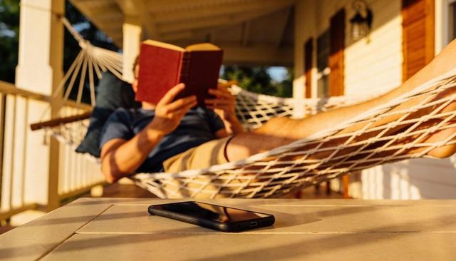 Man reading book in hammock on sunlit porch during golden hour with smartphone in foreground
