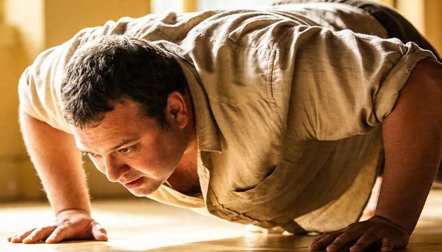 Man performing pushup on polished wooden floor in sunlit interior wearing beige shirt