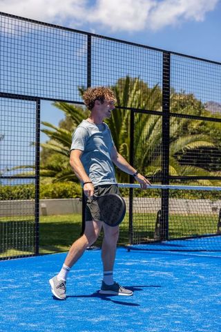 Young man celebrating point on bright blue padel court with tropical greenery