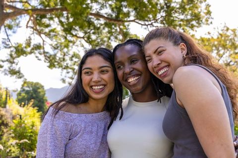 Three diverse women smiling beneath sunlit tree in garden, casual friendship portrait