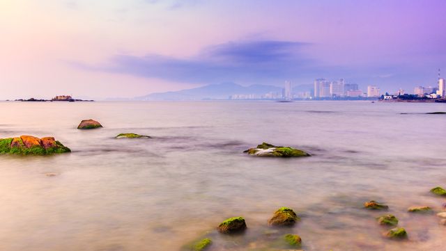 Coastal scene displaying a serene ocean with rocky shore and city skyline under twilight sky. The colorful sunset adds to the tranquil atmosphere, making this ideal for travel magazines, nature websites, and background art focusing on peaceful and calm themes.