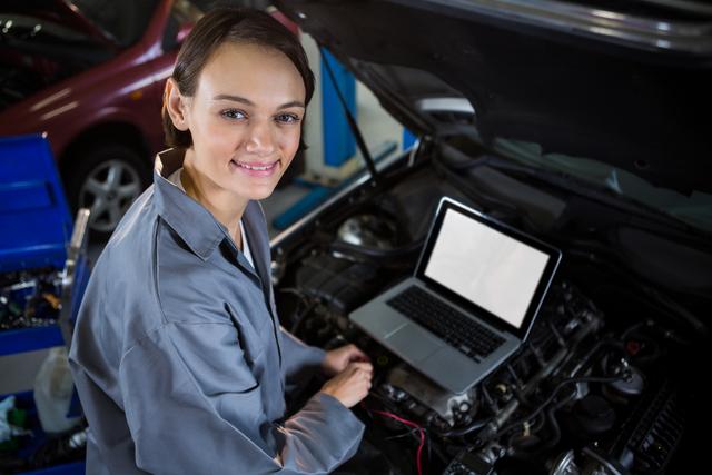 Female mechanic examining car engine with help of laptop at repair garage