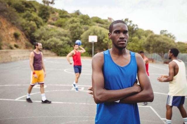 Young male basketball player standing with arms crossed on an outdoor court, exuding confidence. Teammates are seen in the background, engaging in practice. Ideal for use in sports-related content, fitness promotions, teamwork and leadership articles, and advertisements for athletic gear.