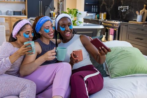 Three african american women enjoying cozy home spa night with under-eye patches and mugs
