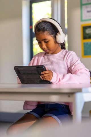 Schoolgirl wearing headphones using tablet at desk focused on digital learning and reading