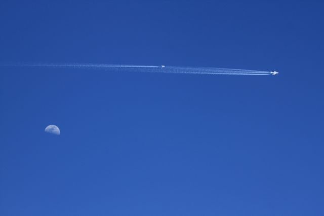 An airplane leaves a white contrail while flying high in a bright blue daytime sky, with the moon visible on the left. This image can illustrate topics such as aviation, clean energy, or NASA research. Suitable for articles, presentations, or educational content focused on aircraft emissions, contrail impacts, and environmental studies.