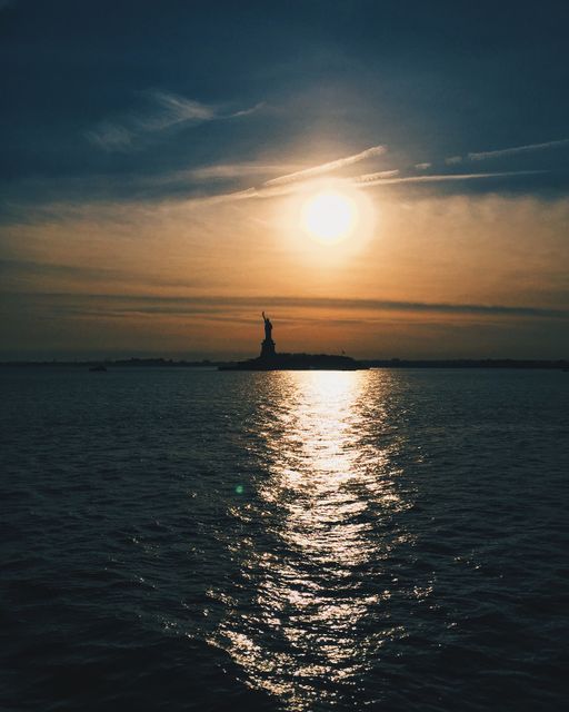 Silhouette of Statue of Liberty at sunset with vibrant sky and reflective ocean water. Ideal for use in travel brochures, websites promoting New York City tourism, inspirational quotes, or backgrounds for presentations highlighting iconic landmarks.
