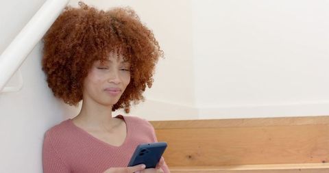 Woman sitting on stairs leaning on railing using smartphone in sunlit minimalist home