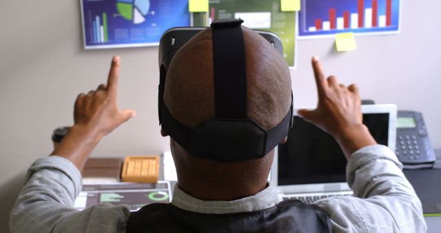 Shot of a businessman facing computer while wearing a VR headset, interacting with virtual graphs and charts. Ideal for illustrating advancements in technology, innovative business practices, or modern office environments. Great visual for tech blogs, business articles, or presentations on futuristic workplace tools.