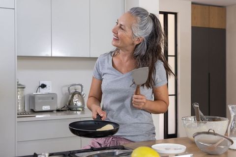 Senior woman cooking pancakes at modern kitchen island, smiling and flipping with spatula